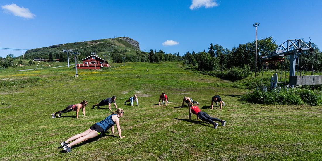 En gruppe mennesker som tar pushups i herlige omgivelser på en grønn gressplen med fjellet Skeikampen i bakgrunnen. 