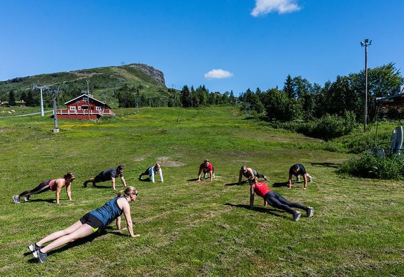 En gruppe mennesker som tar pushups i herlige omgivelser på en grønn gressplen med fjellet Skeikampen i bakgrunnen. 