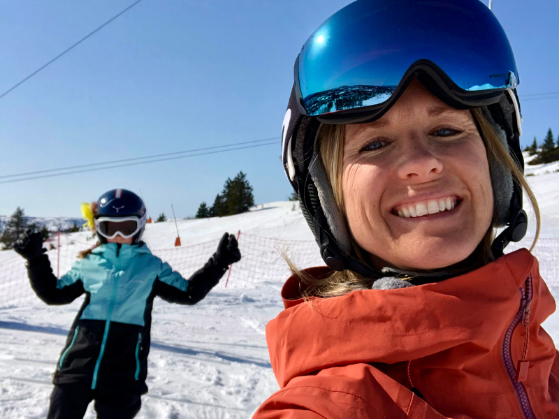 Voksen kvinne som smiler med alpinhjelm og skibriller, jente i bakgrunnen